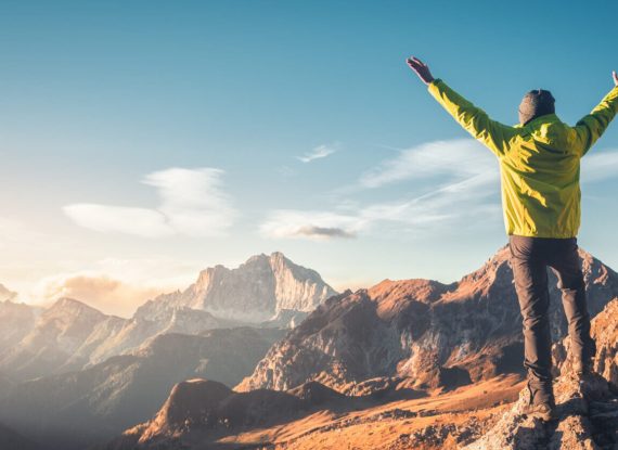 Sporty man standing on the stone with raised up arms against mountain valley at sunset. Happy young man, rocks, autumn forest and blue sky in Dolomites, Italy. Traveler hiking in alpine mountains